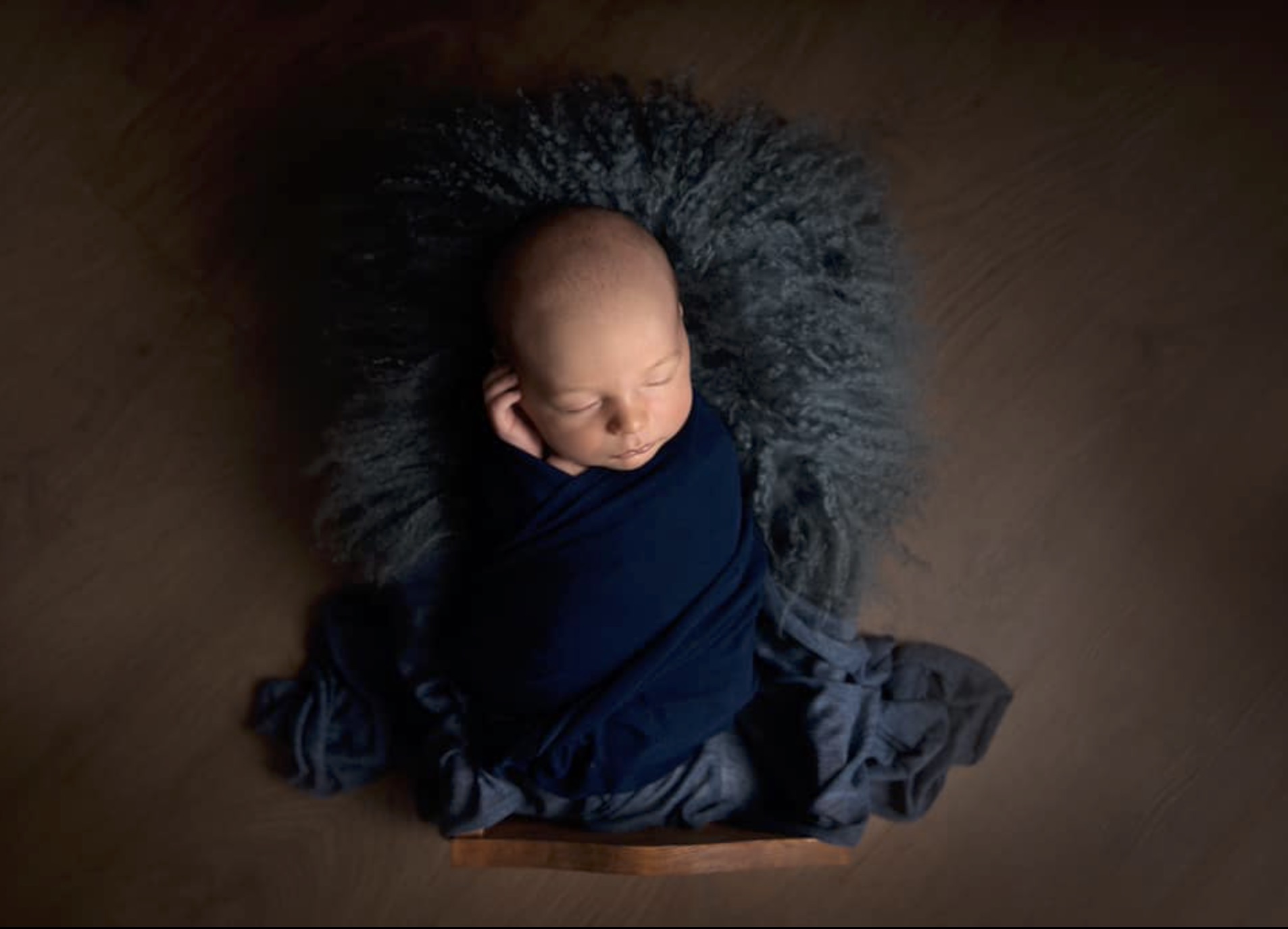 newborn baby boy wrapped in navy blue wrap posed in a little wooden bed on a blue fur rug