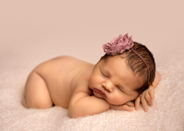 Newborn baby girl wearing a dusky pink floral headband, sleeping on her tummy on a pink blanket, with her head resting on her hands.