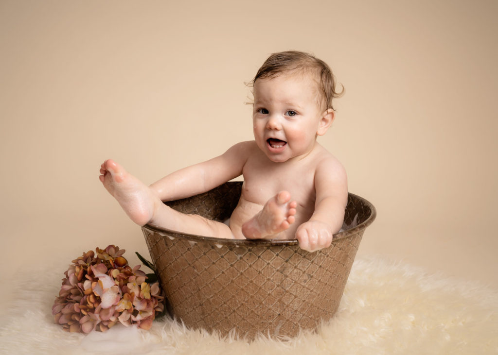 Smiling one year old sitting in a vintage bath tub on a cream fur rug, with legs poking out.