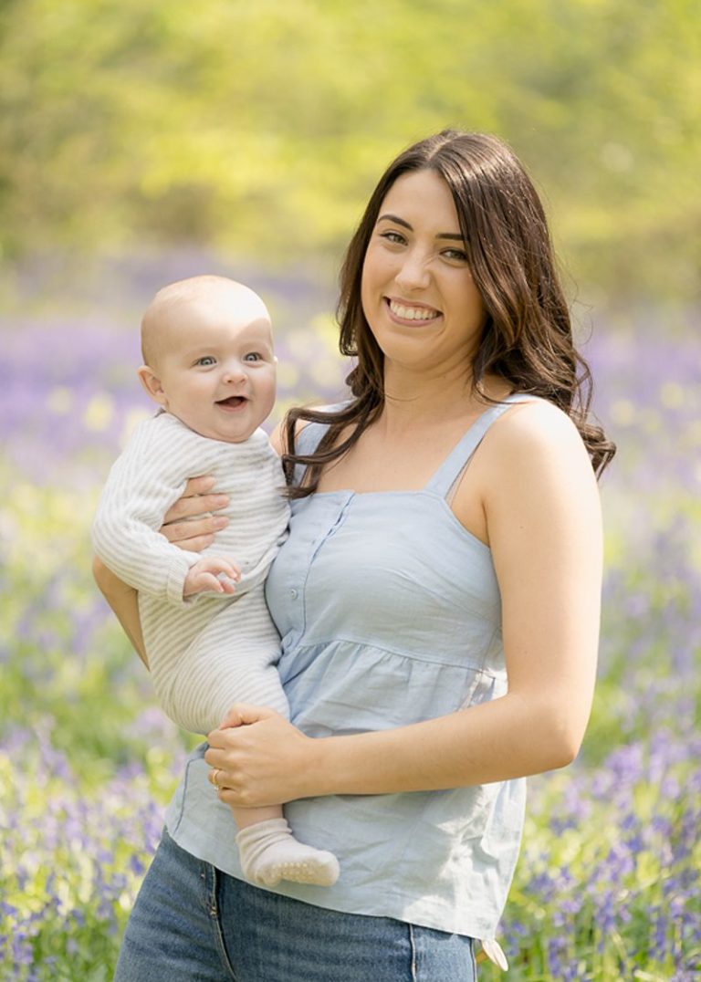 Young mum with long dark hair, wearing a pale blue vest top, holding her baby in the bluebell woods.