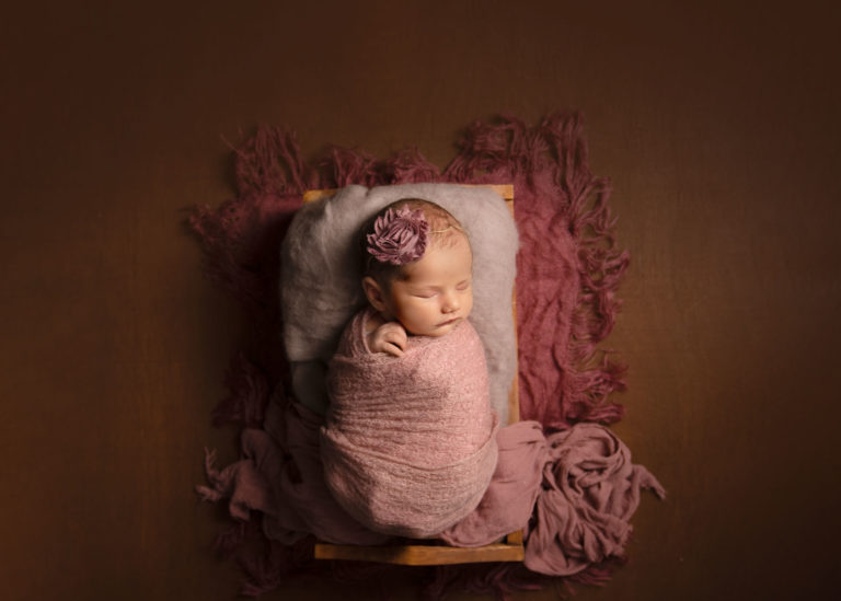 Newborn baby girl in a soft pink wrap, with a pink flower in her hair, sleeping in a small bed, surrounded by soft pink blankets, by newborn Photographer Andover.