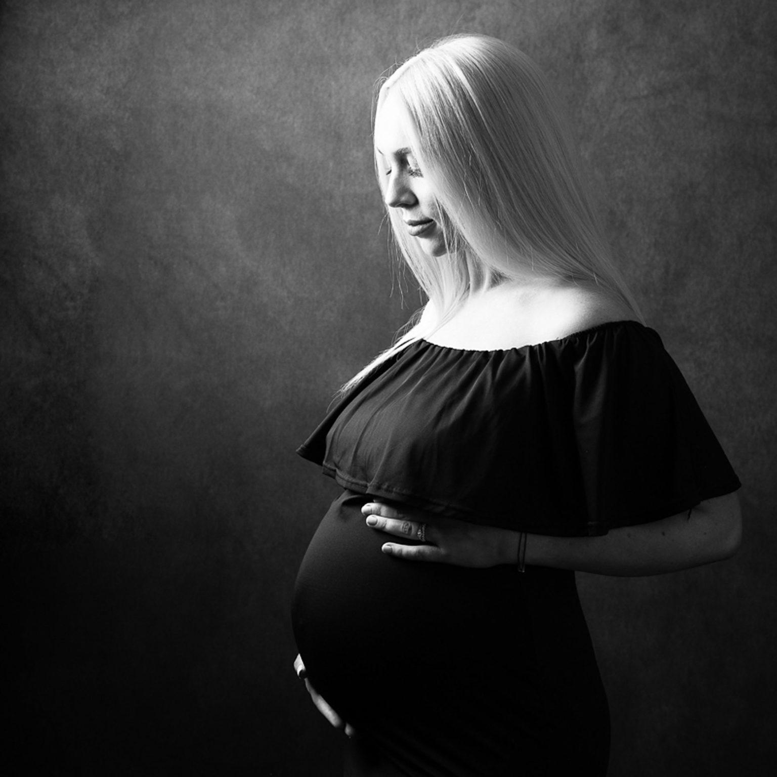 Pregnant lady wearing a white sheer dress, with bump exposed, smiling at the camera against a studio backlit white.