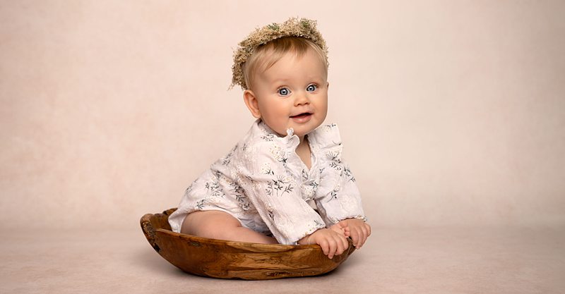 A young girl wearing a white floral romper suit and a floral headdress, sitting in a small wooden bowl, smiling at the camera.