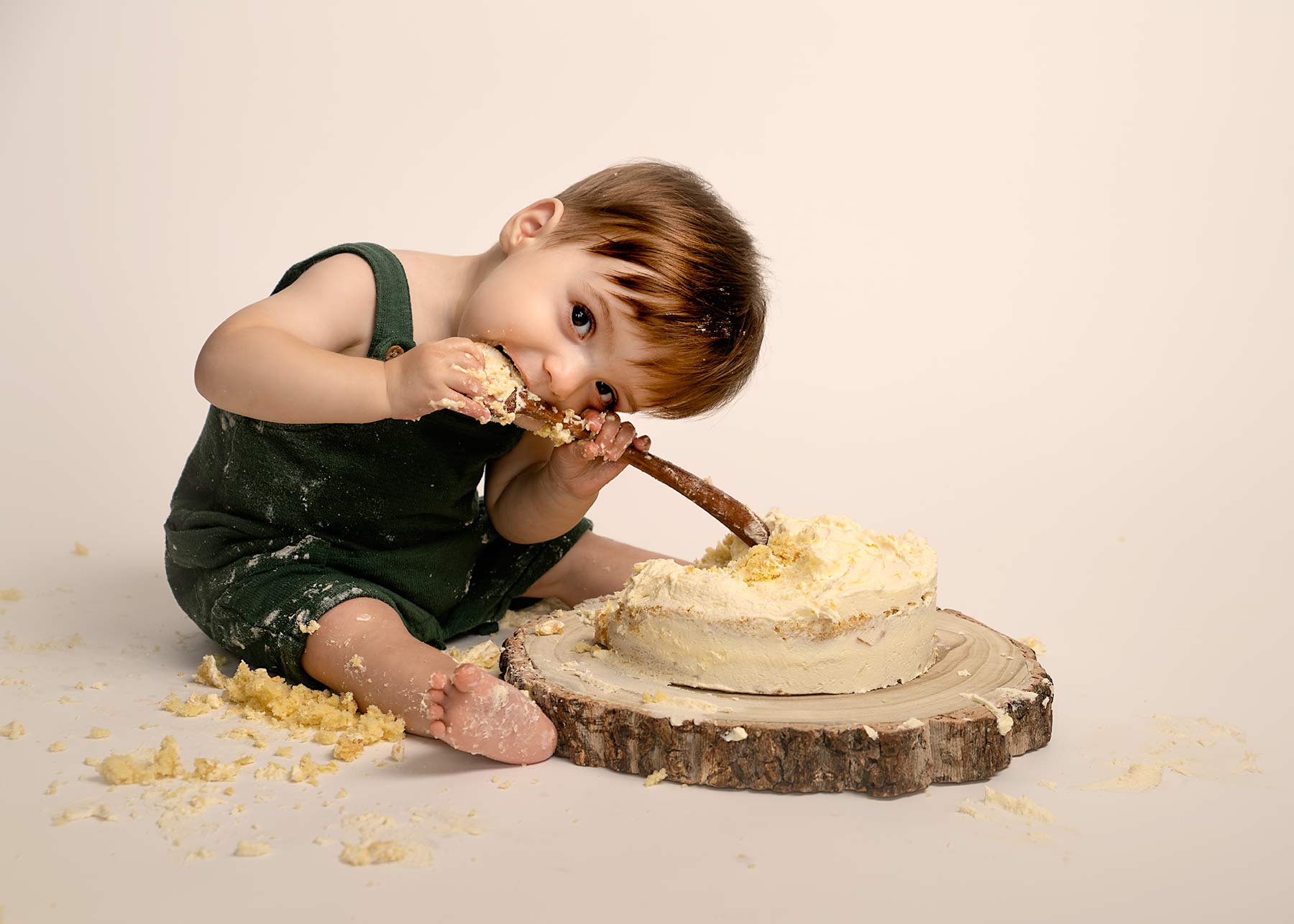 One year old boy in blue romper suit smiling at camera in front of a cake he is about to smash with a wooden spoon.