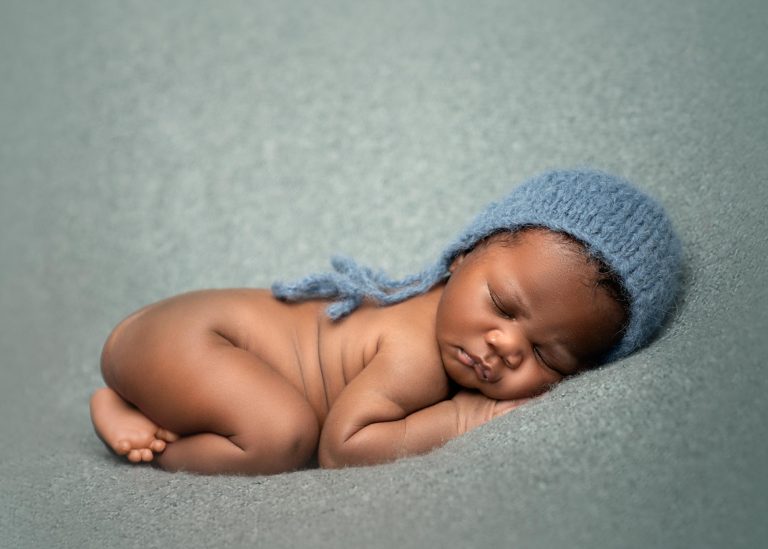 Newborn baby with black skin and blue mohair hat, sleeping on his front on a blue blanket.