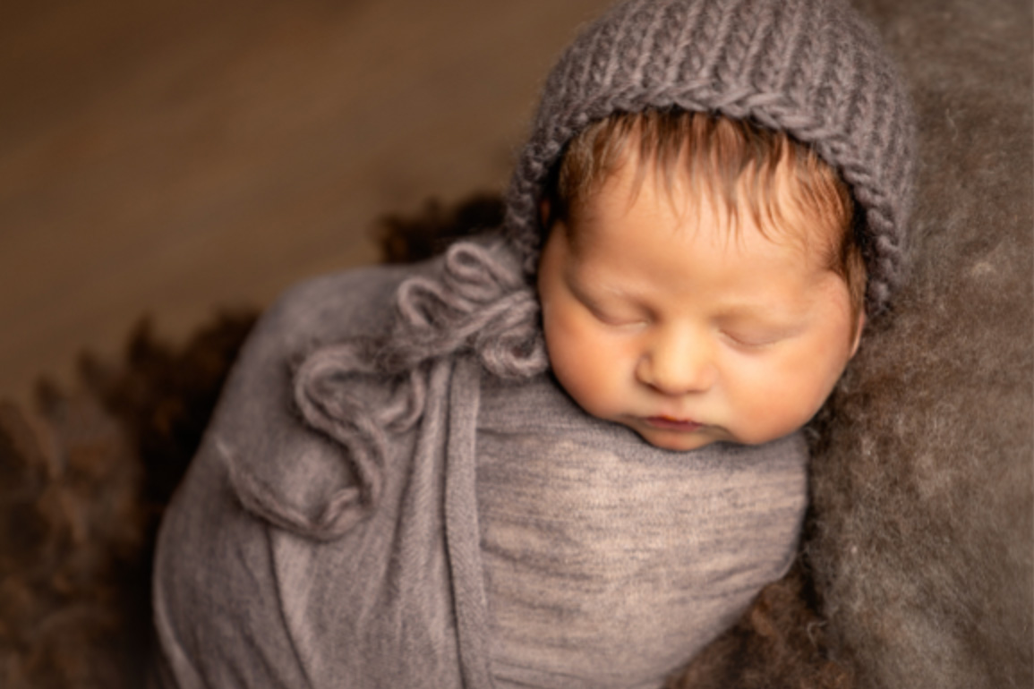 Newborn baby swaddled in a soft brown wrap with a knitted bonnet, sleeping in a little log bed.