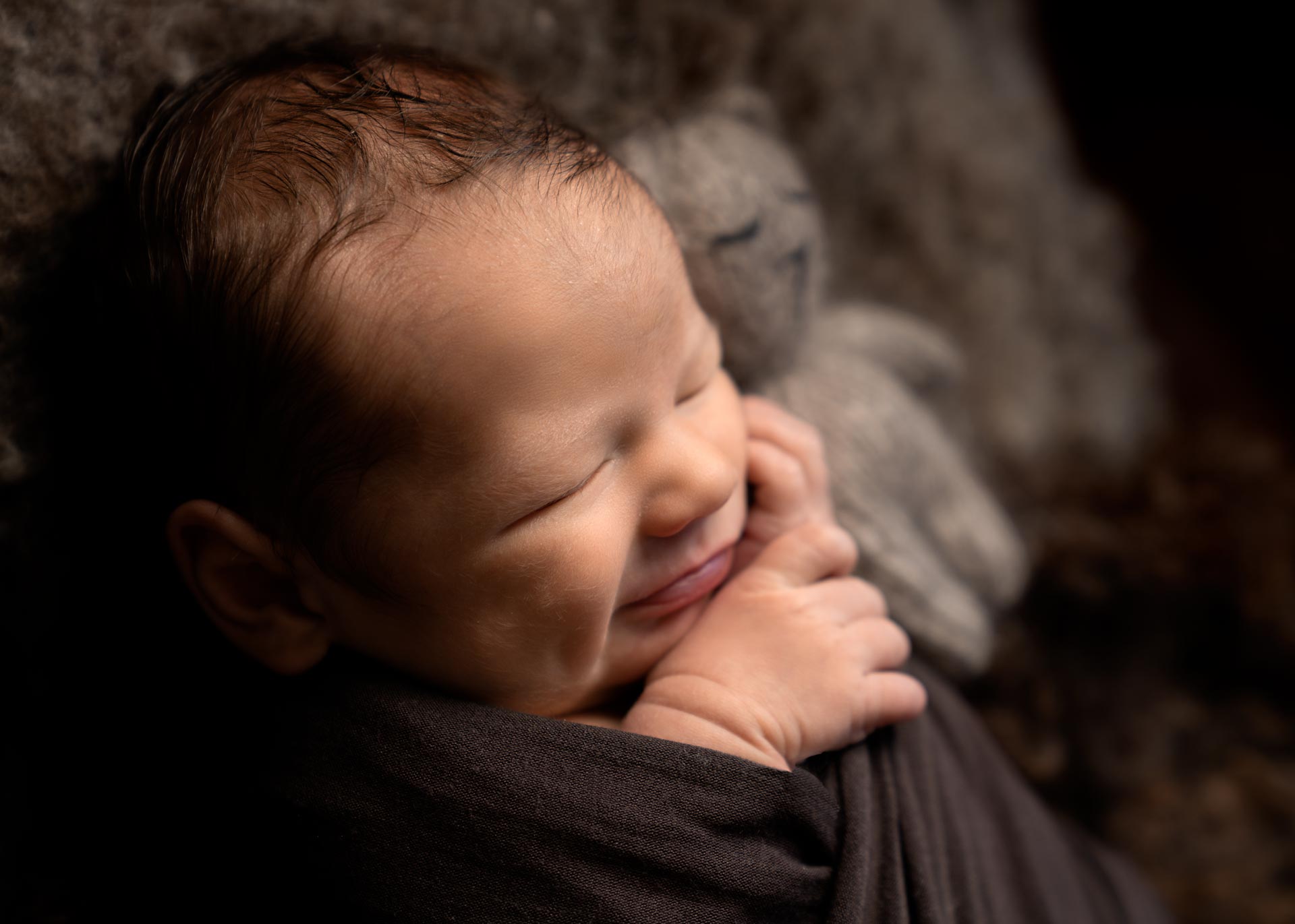 newborn baby smiling whilst swaddled in a brown wrap next to a knitted teddy bear