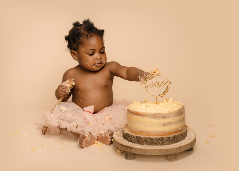 One year old birthday photoshoot image of young girl wearing an oversized pink tutu, about to smash a cake.