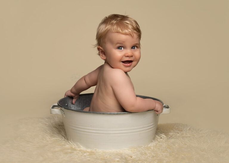 One year old boy sitting in a vintage style bath tub, on a white fur rug, cleaning up at the end of a cake smash photoshoot.