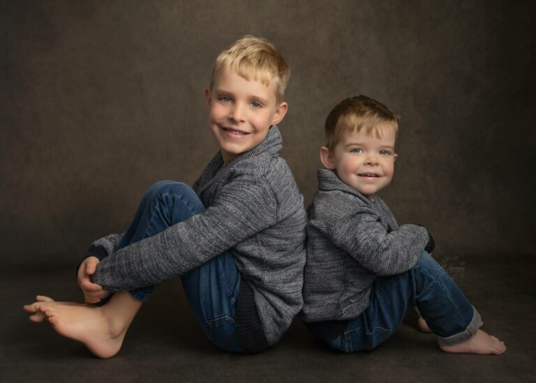Portrait of two young brothers, wearing matching knitted jumpers and jeans, sitting back to back on a dark brown backdrop.