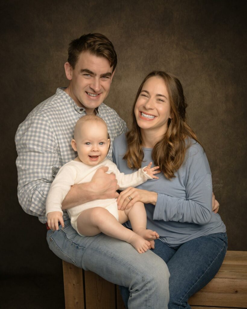 Family portrait of young couple with their baby, sitting at an Andover photography studio against a dark backdrop.