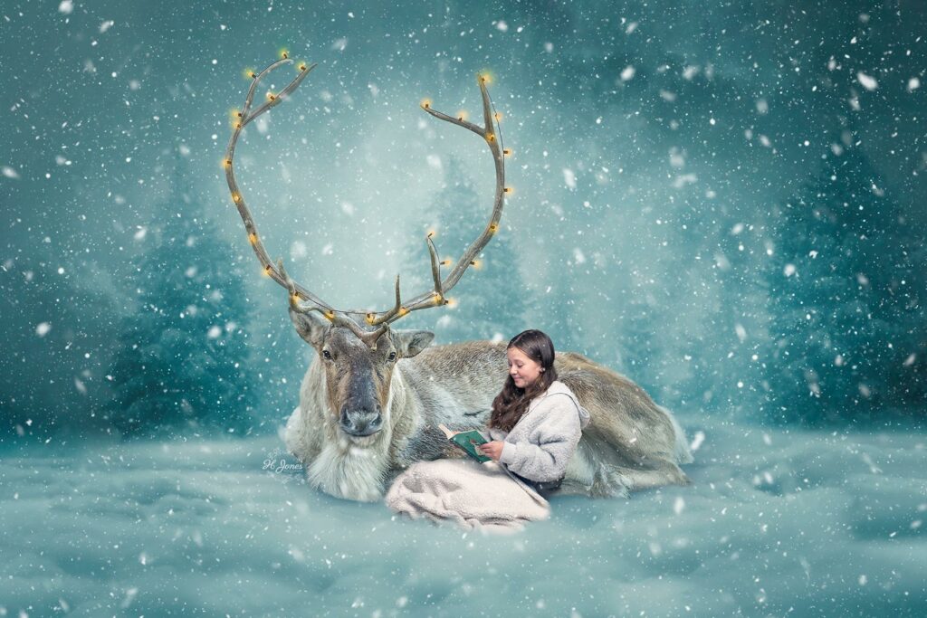 Girl snuggled under a blanket, reading a book, leaning against a reindeer lying in the snow.