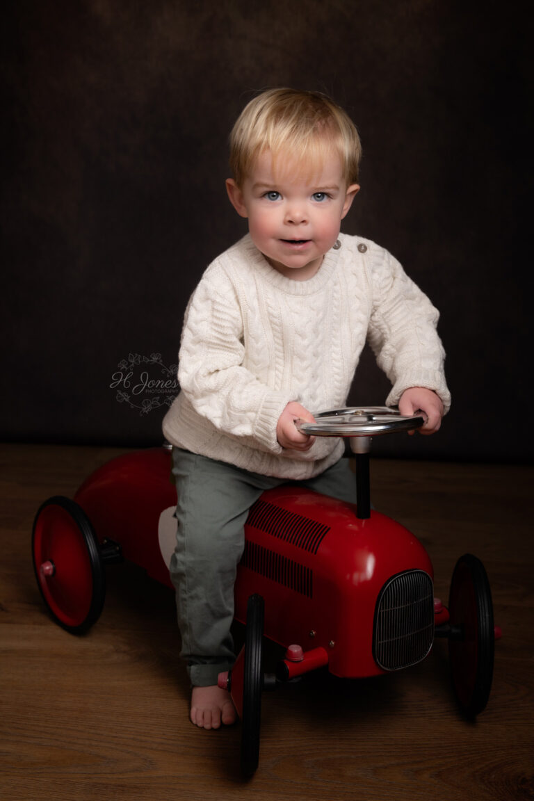 Portrait of small boy sitting on red toy car, wearing a chunky white jumper.