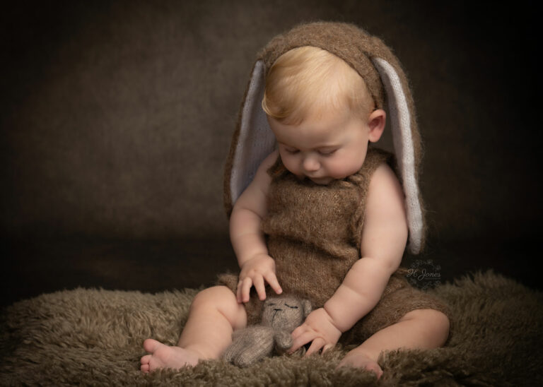 Portrait of small boy in brown fluffy romper with bunny ears, playing with small teddy bear, sitting on bon fur rug.