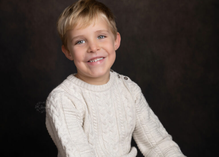 Portrait of boy in white jumper, smiling at the camera, against a dark backdrop.