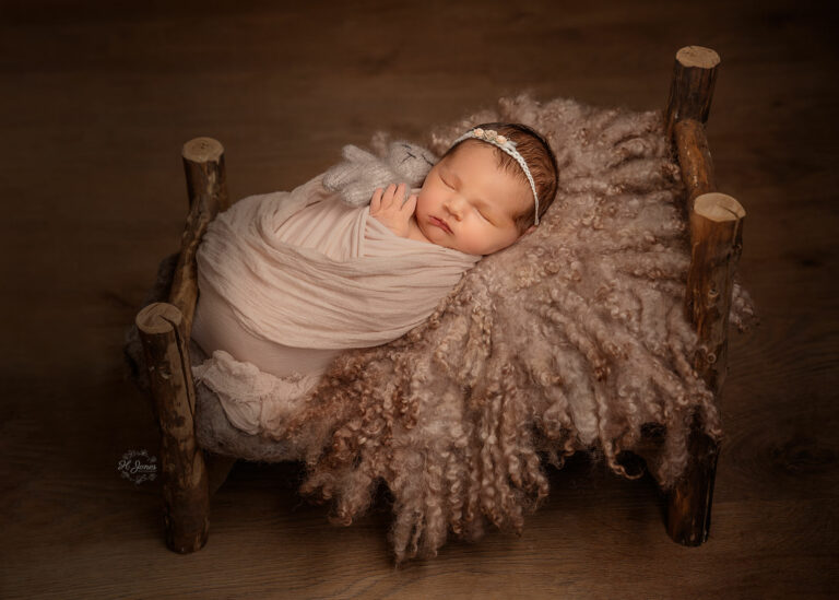 Newborn baby girl wrapped in dusky pink wrap, wearing a white headband, lying in a small wooden bed with fur blanket.
