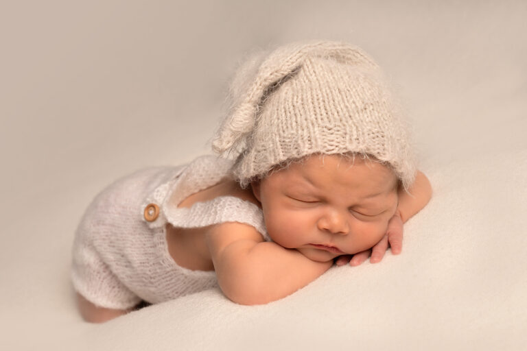 Newborn baby sleeping. on its front, with head resting on arms, wearing a knitted hat and outfit on a cream blanket.