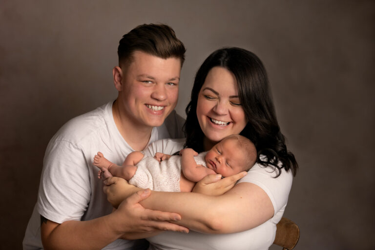 A young couple wearing white t-shirts, laughing and holding their newborn baby, in a photography studio.