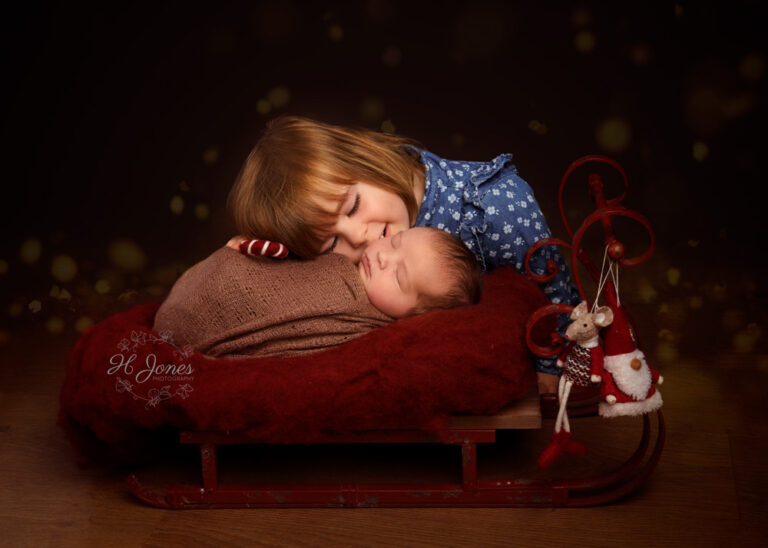 Young girl wearing a blue dress cuddling her baby, lying on a miniature Christmas sledge, on red fur.
