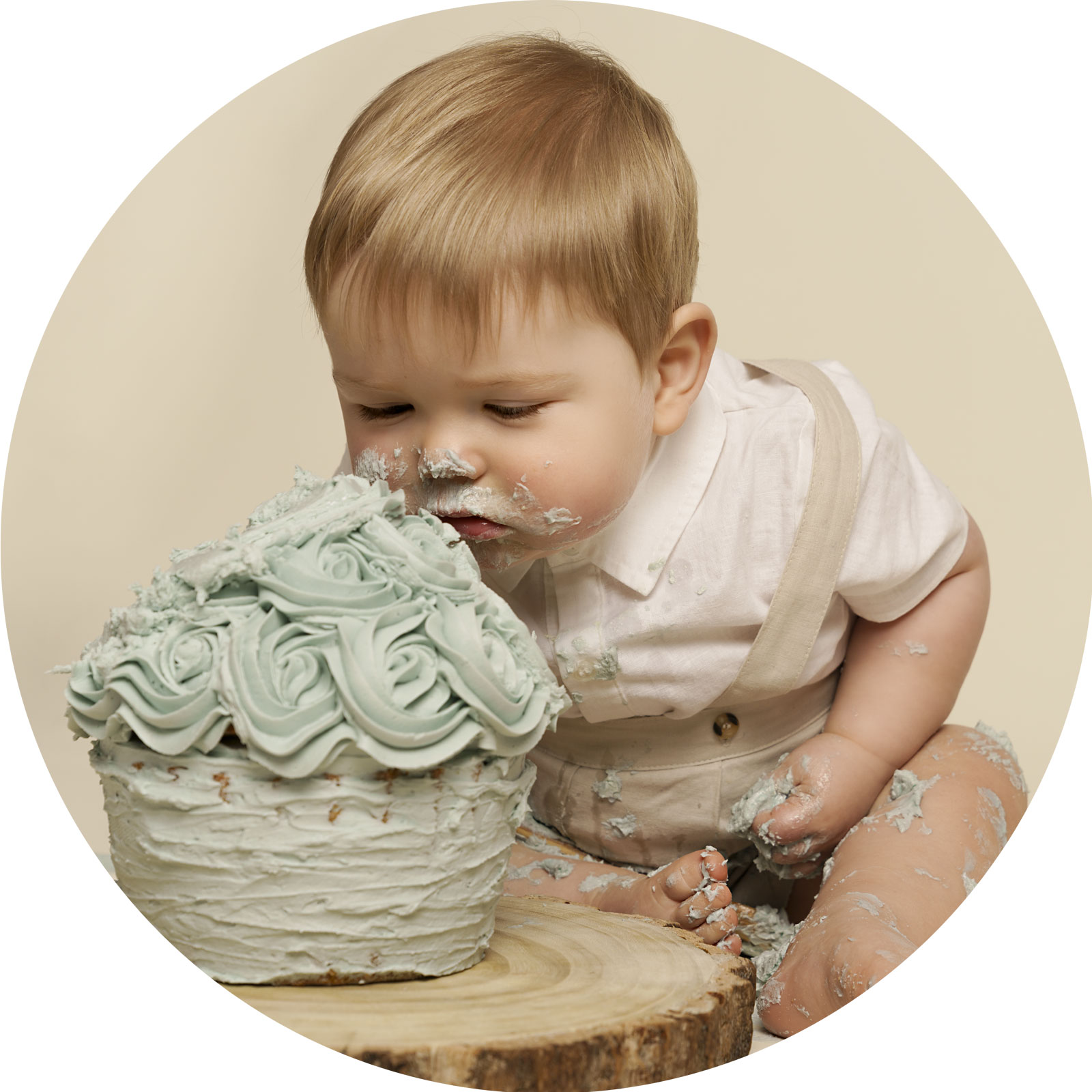 One year old boy dressed in a white shirt and cream coloured dungarees, planting his face into a blue coloured giant cupcake.