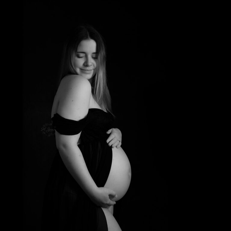 Pregnant woman posing in a black dress, with her bum on show, against a dark backdrop.