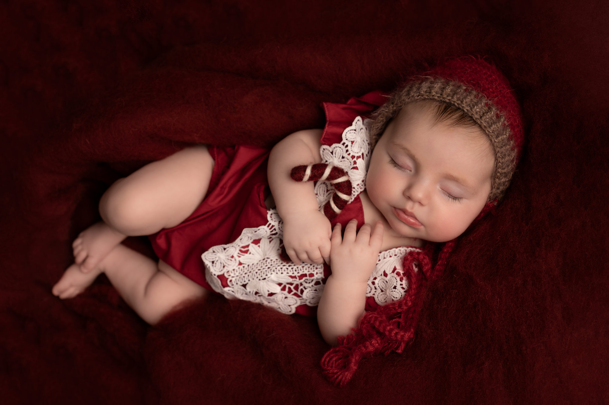 Baby girl dressed in a red Christmas outfit sleeping on a red fur blanket, cuddling a knitted candy cane.