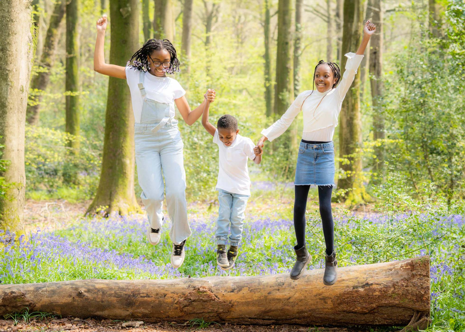 3 siblings holding hands jumping off a log in the Hampshire bluebell woods.