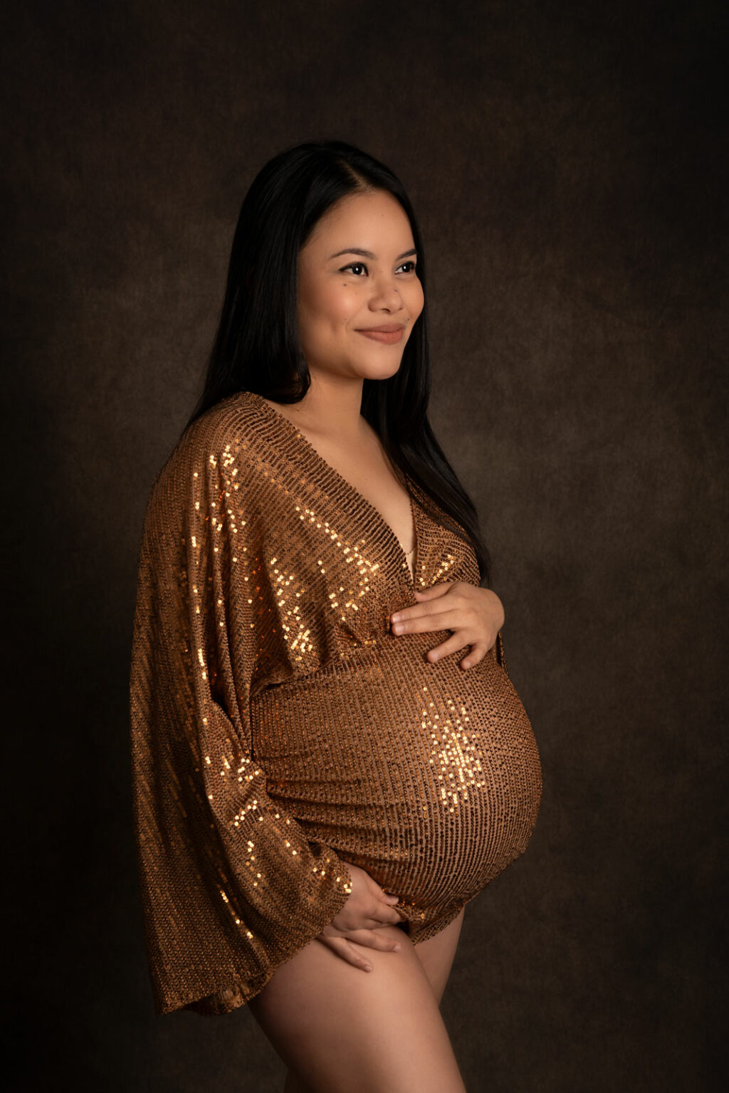 Pregnant lady posing in a gold glittery bodysuit against a dark backdrop for a maternity photoshoot.