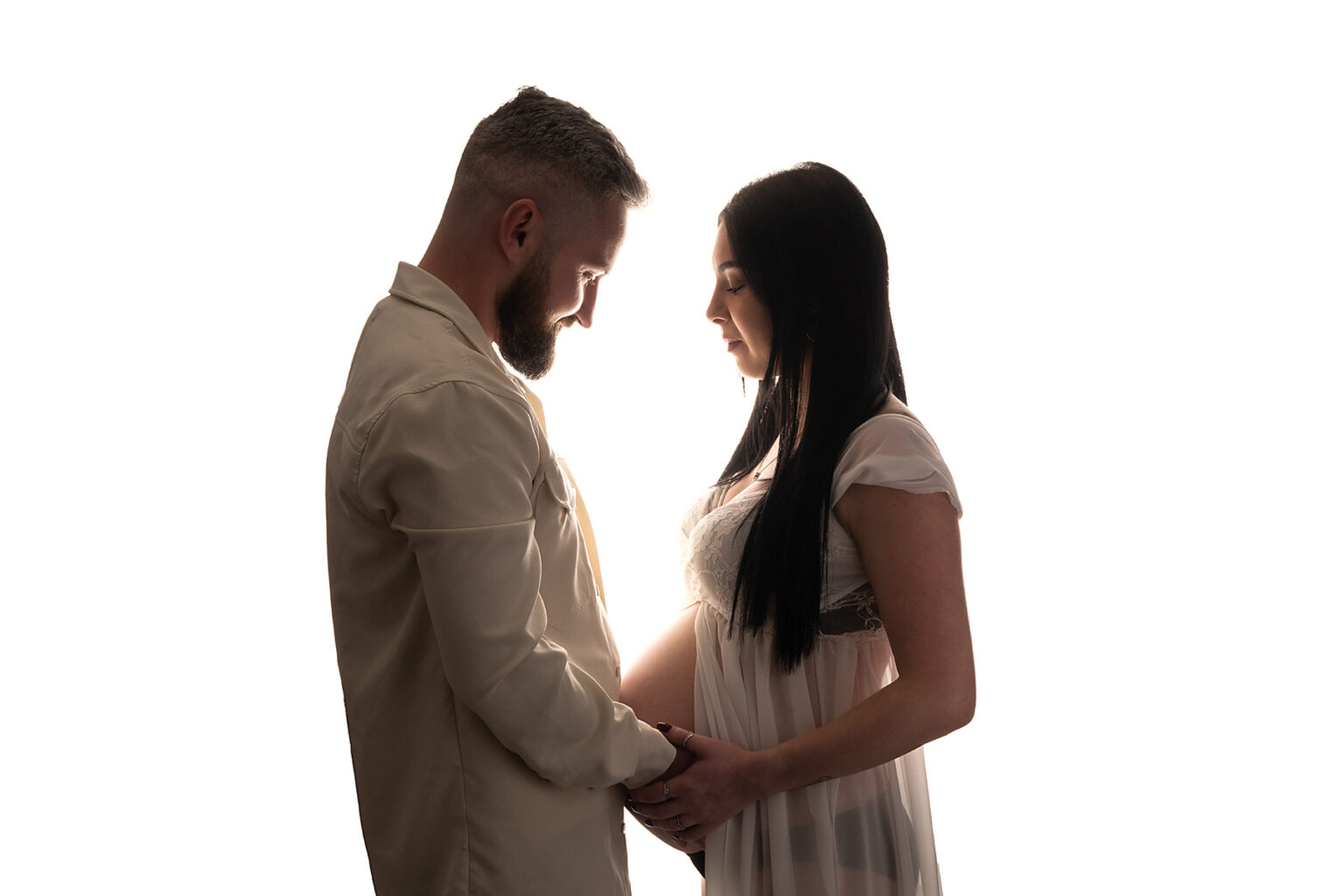 Pregnant couple facing each other, dressed in white, with bump exposed, gazing down at the bump.