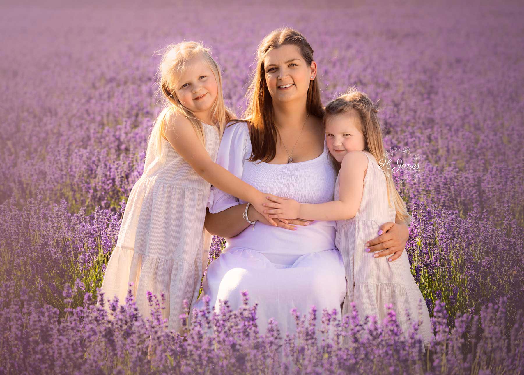 Pregnant mother and two daughters, all wearing white dresses, posing for an outdoor maternity photoshoot in lavender fields of Hampshire.