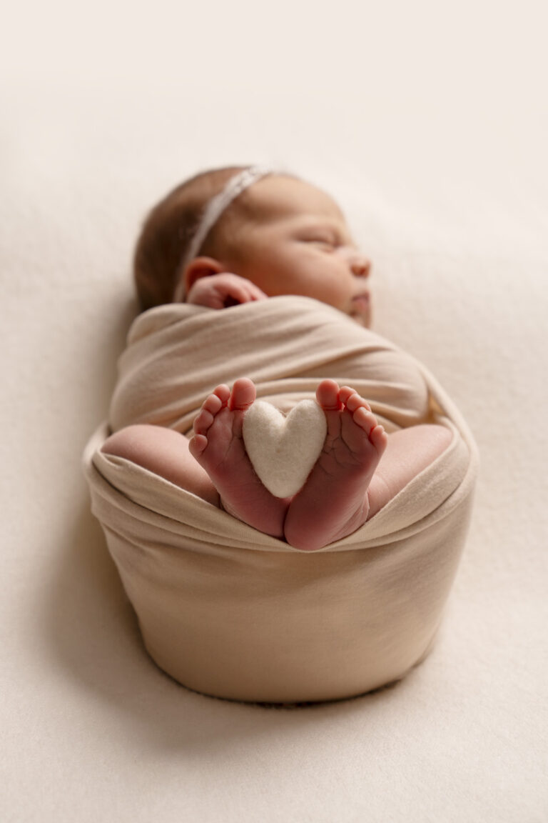 Newborn baby girl, wrapped in a pale pink swaddle, with feet exposed, holding between them a small knitted heart.