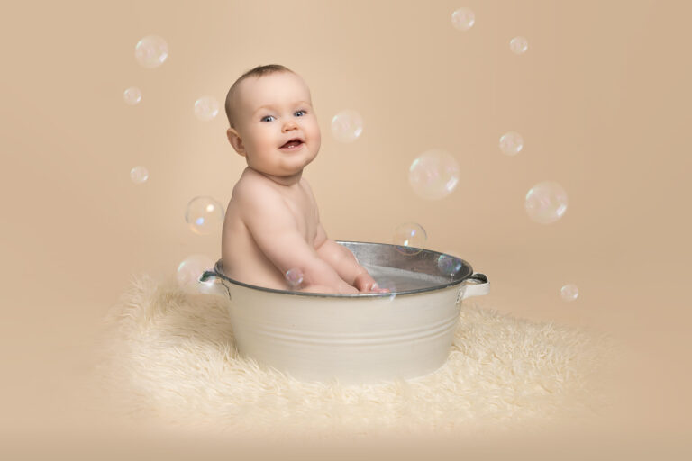 One year old boy in vintage style tub, smiling at camera at cake smash and splash photoshoot, surrounded by bubbles.