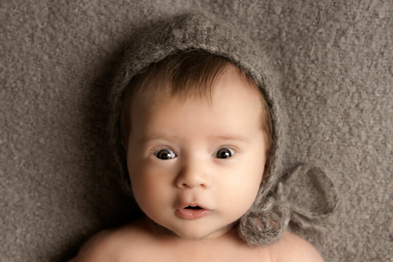 Close up of baby wearing a knitted grey bonnet, lying on a grey blanket, looking directly at the camera.