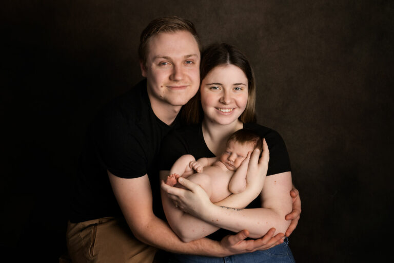 Young couple posed in black, against a black backdrop in a photography studio, Hampshire, cradling their newborn baby, smiling.