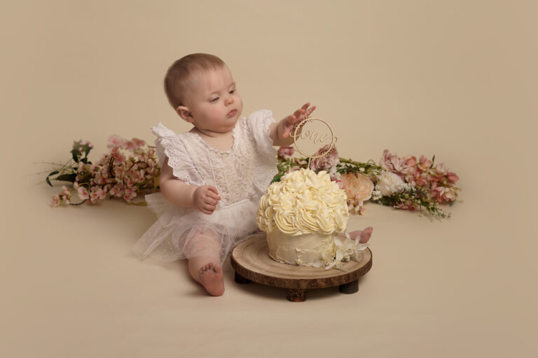 One year old girl in a white lace romper suit, sitting behind a giant cream cupcake, with flowers behind her, at a cake smash photoshoot in Hampshire.