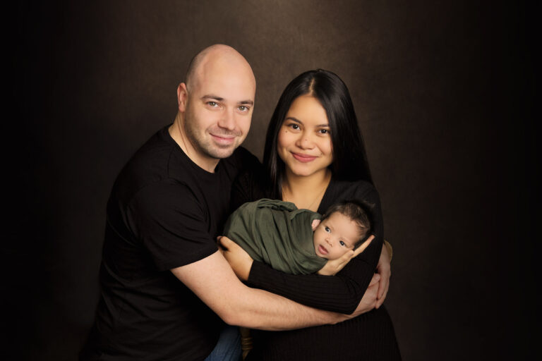 Couple wearing black, holding their newborn baby wrapped in a dark green swaddle, at H Jones Photography studio in Hampshire.