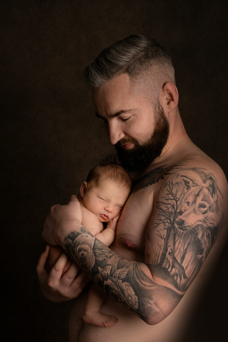 New dad posing without top, showing his tattoos, cradling his newborn baby.