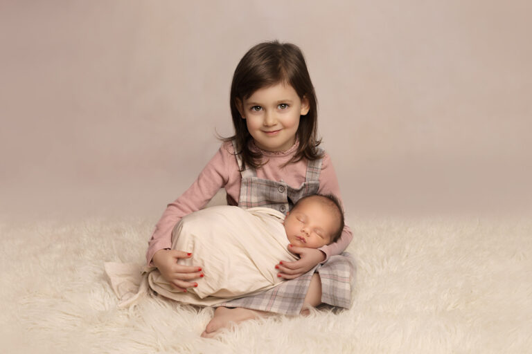 Proud two year old sister wearing a pink t-shirt and grey checked dress, sitting cross legged on a white fur rug, cradling her newborn baby brother, wrapped in a white swaddle.