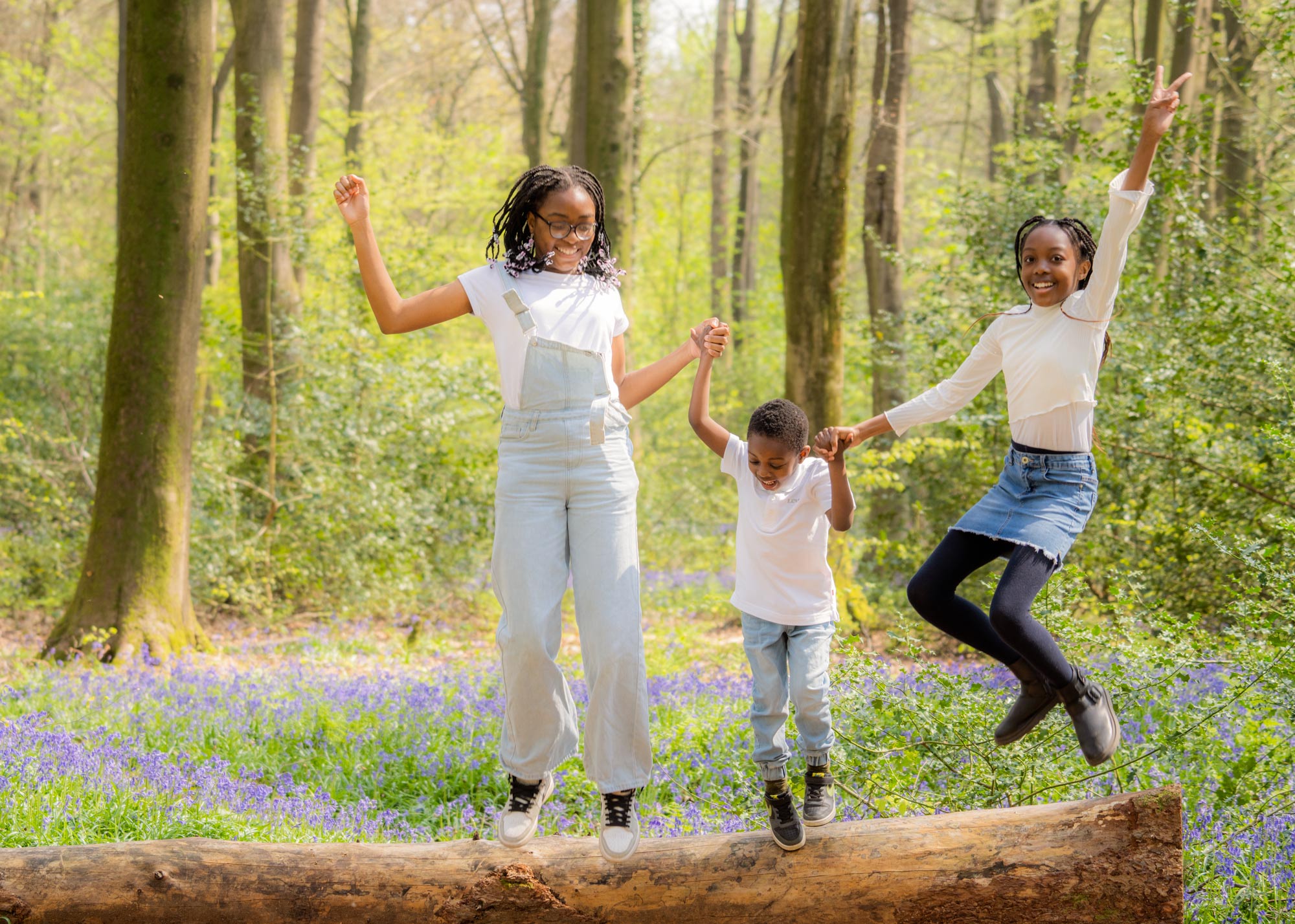Three children wearing white t-shirts, jumping off a fallen tree in the bluebell woods, smiling.