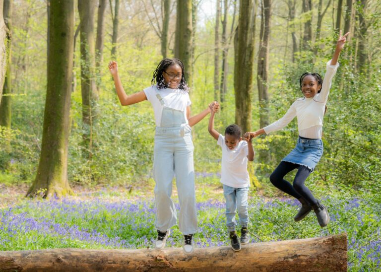 Three children jumping off a fallen log in the bluebells at a family photoshoot in Hampshire