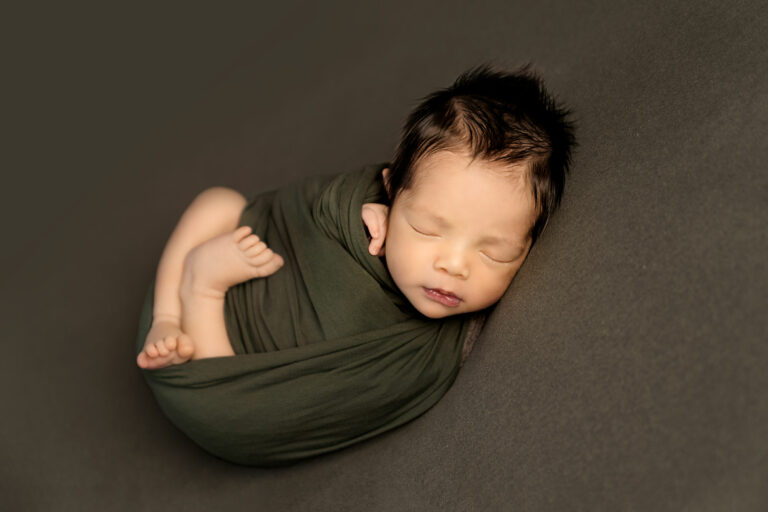 Newborn baby boy curled up in a dark green wrap, sleeping posed at H Jones Photography studio in Hampshire.