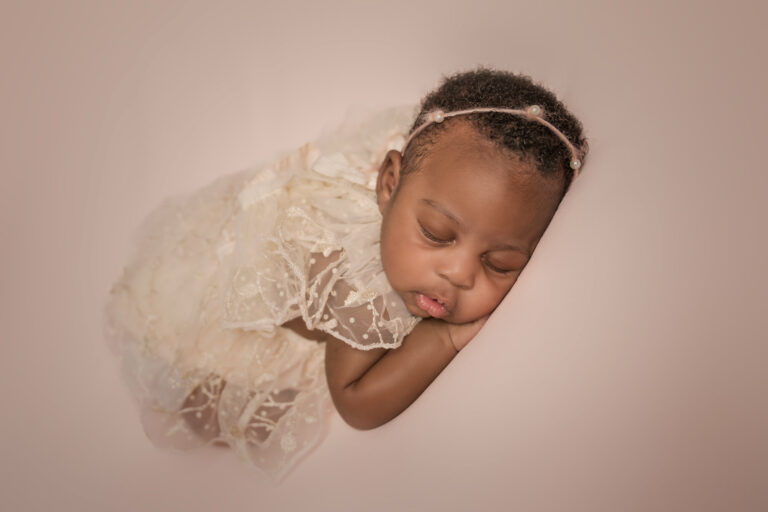 Newborn baby girl in a cream coloured lace outfit, with a pink perl headband, sleeping on her front at H Jones Photography studio in Hampshire.