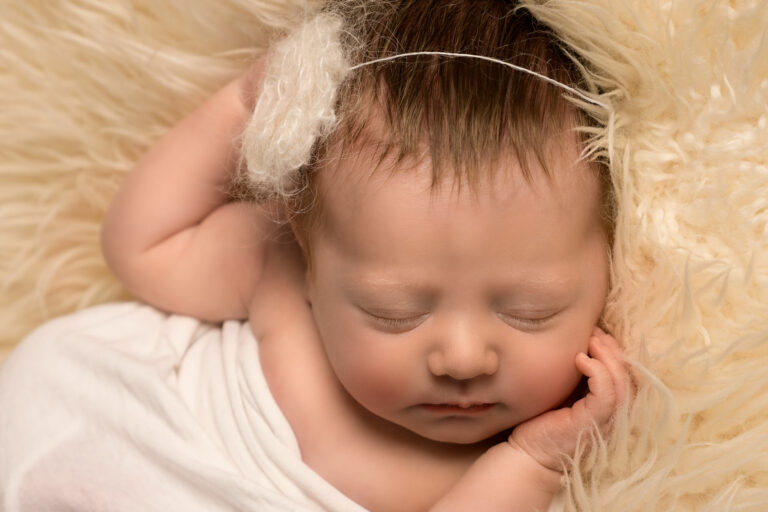 Close up of newborn baby girl, wearing a white mohair flower headband, holding one hand to her face, sleeping on a white fur rug.