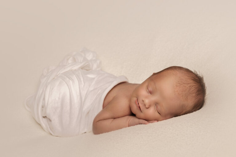 Newborn baby swaddled in a white wrap, sleeping on their front, on a white blanket, sweetly smiling.