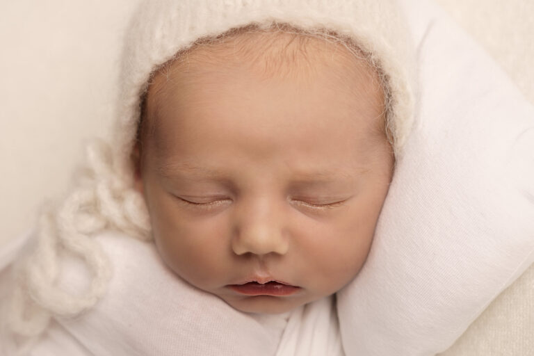 Close up of a newborn baby's face, sleeping on a white blanket, wearing a white knitted hat.