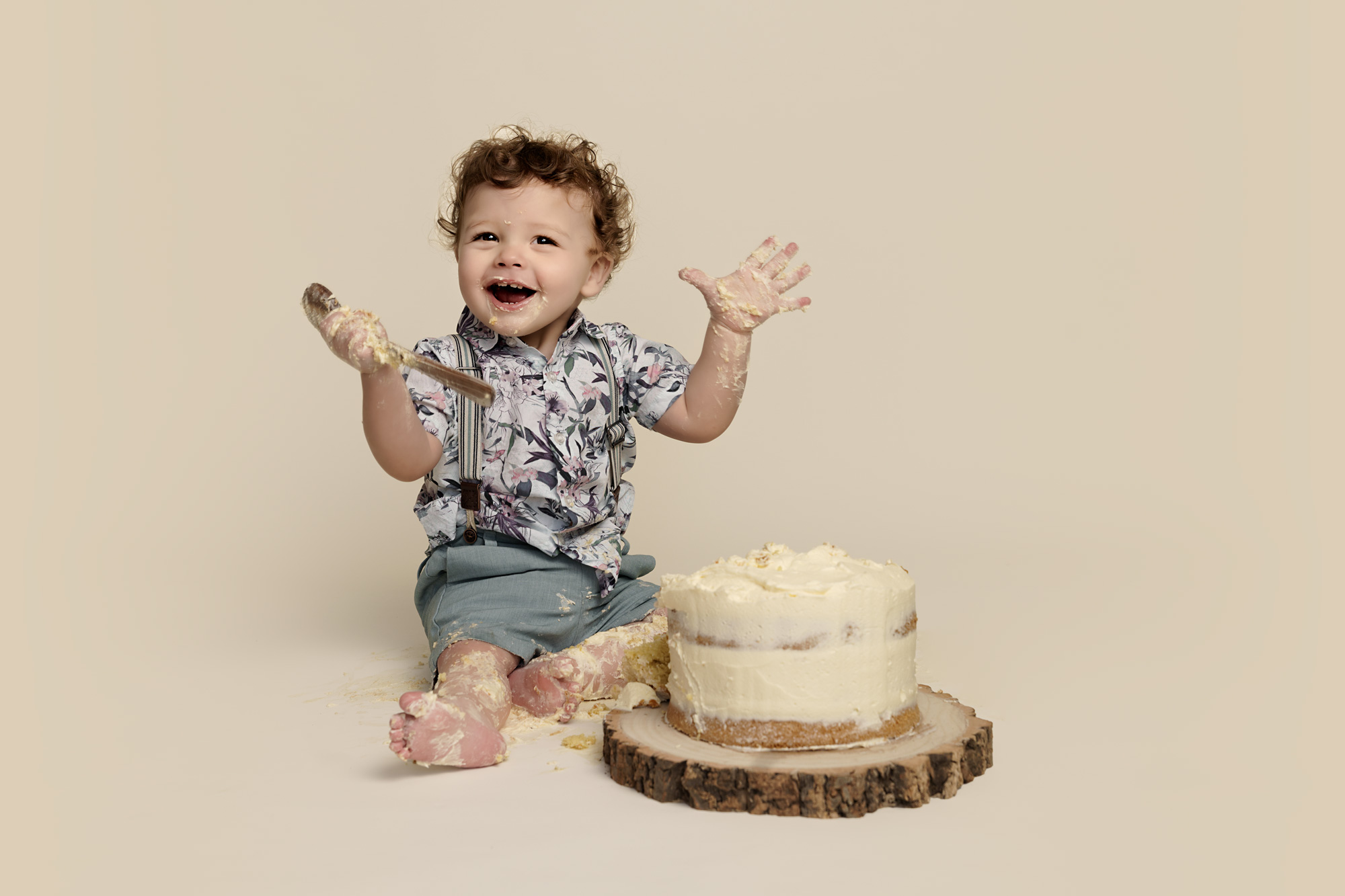 One year old boy sitting gin front of a cream coloured cake, holding a wooden spoon, with icing on his hands and feet, smiling at the camera.