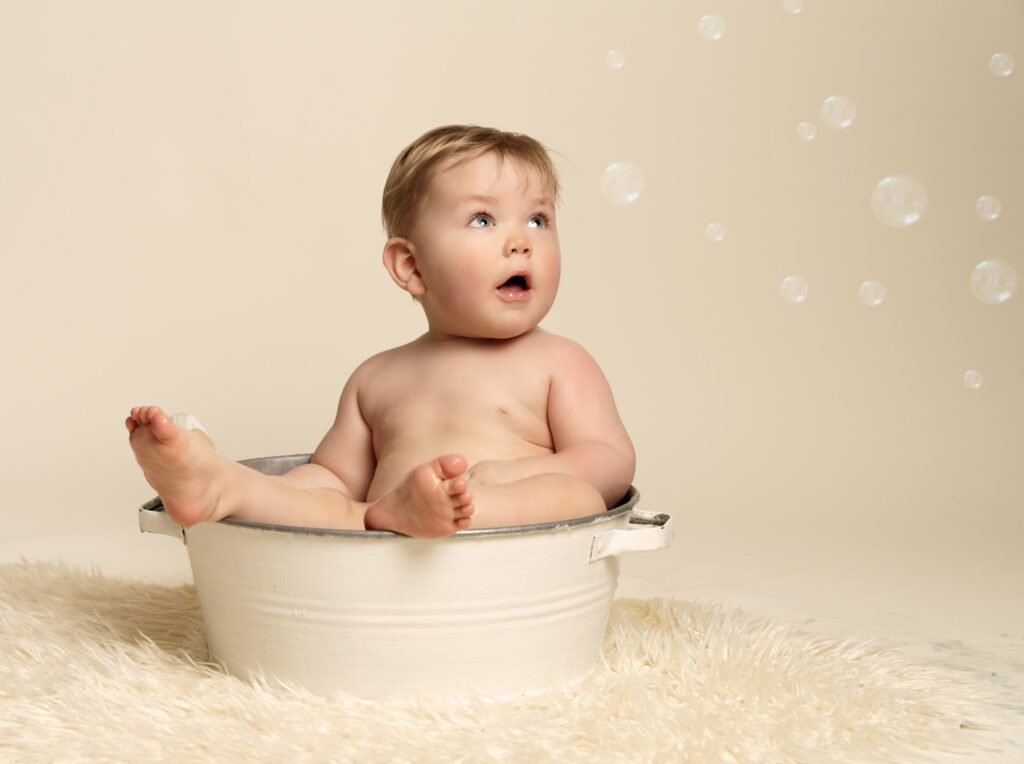 One year old baby boy sitting in a vintage style white bath tub, feet sticking out, looking wide eyed and mouth opened at bubbles falling nearby.