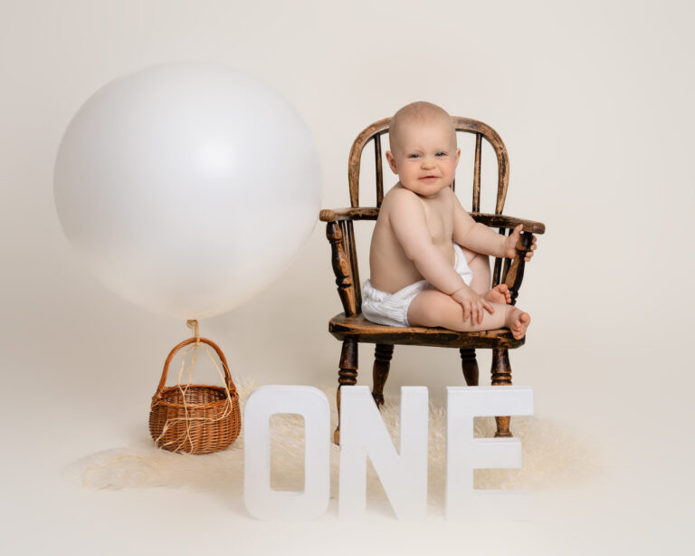 A one year old boy, wearing white shorts, sitting in a vintage child's wooden chair, with an oversized balloon and mini basket, letters spelling the word ONE grinning at the camera.