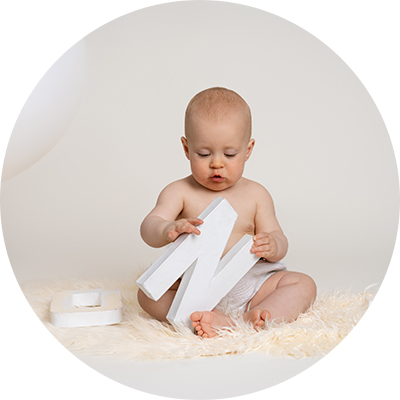 One year old boy, wearing white shorts, sitting on a cream fur rug, looking down at a wooden letter N that he's playing with.