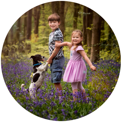 Two young children playing in the bluebell woods with their dog, looking at the camera and smiling.